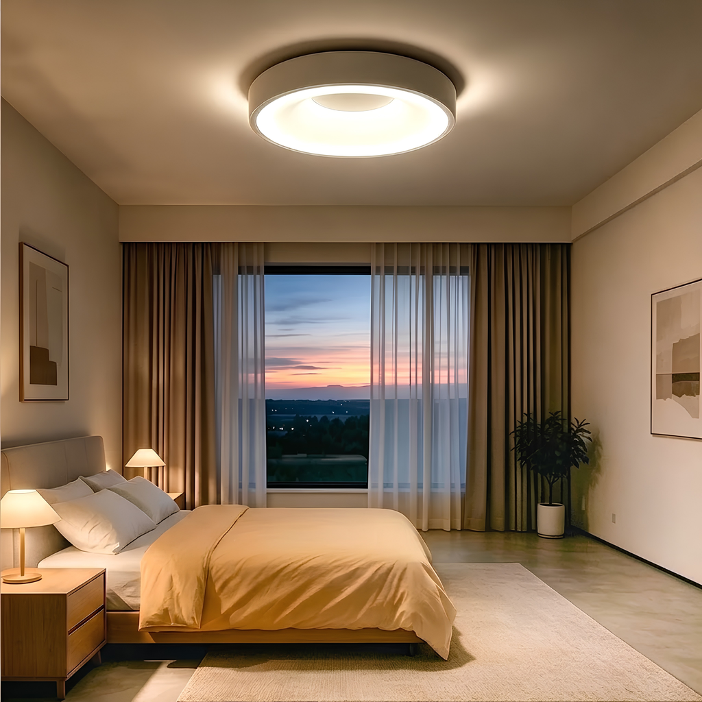 Matte white LED flush mount drum ceiling light glowing warmly above a bed at dusk.