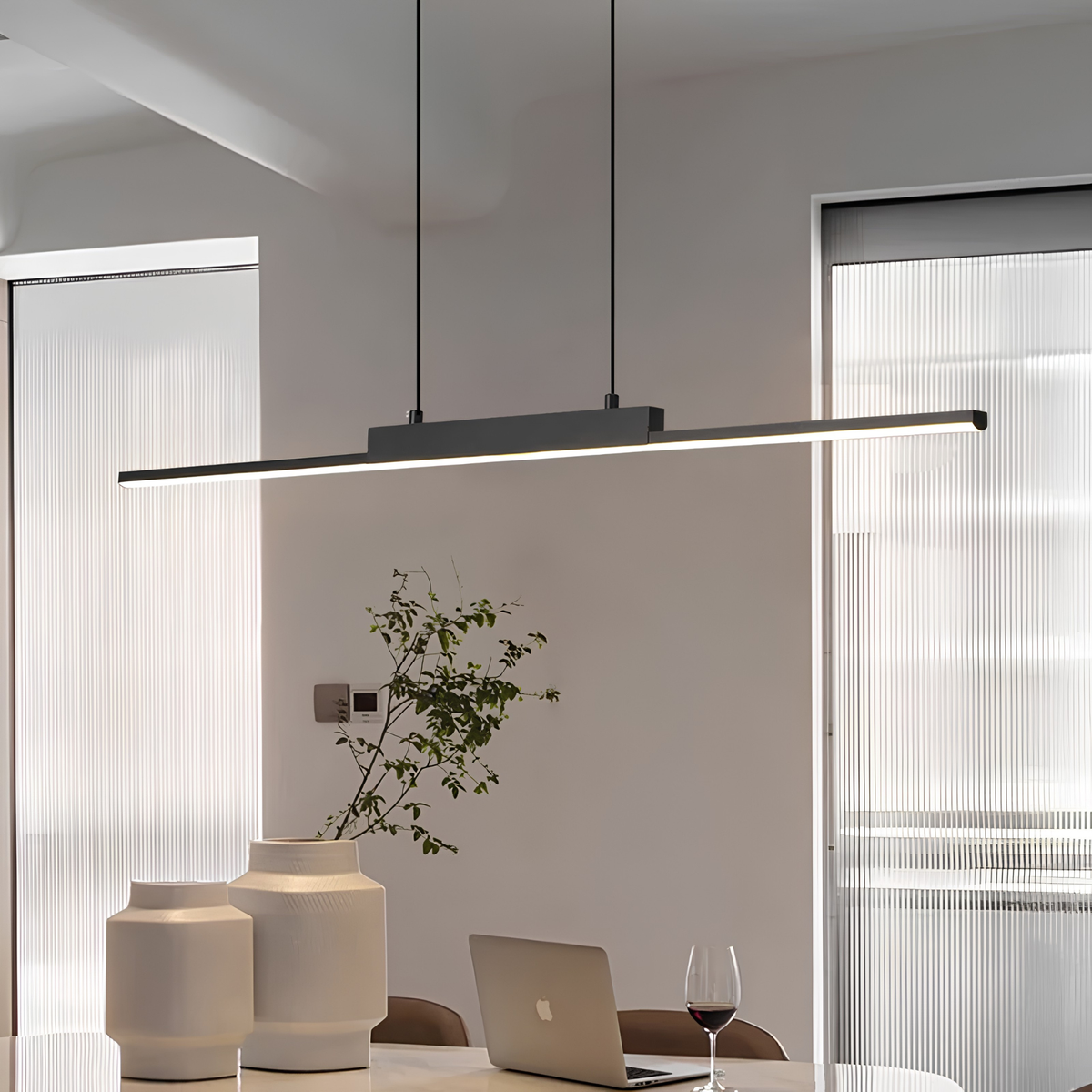 Matte black linear LED pendant casting a warm 3000K glow over a marble dining table set with a laptop and glass of red wine in a minimalist interior.