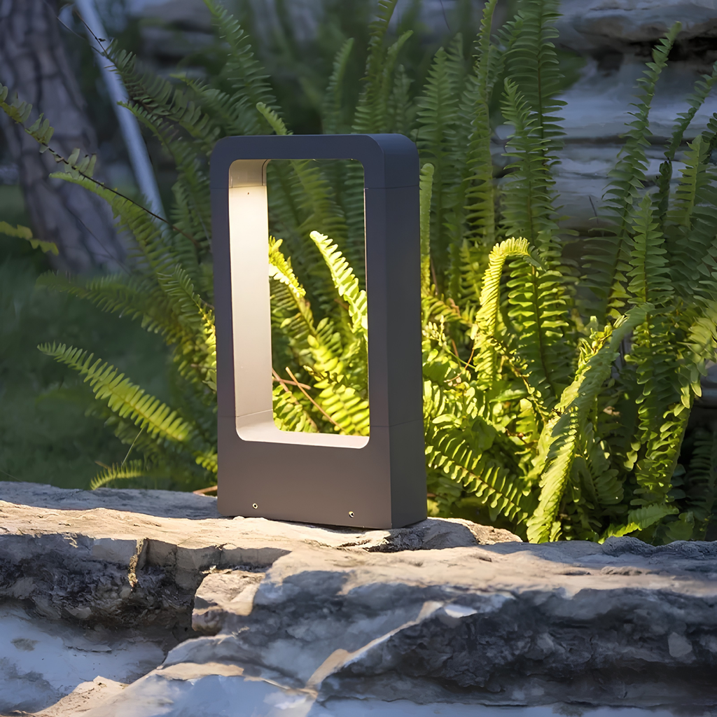 Matte black LED pathway bollard light glowing with a warm 3000K light amid fern foliage on a sandstone garden ledge at dusk.